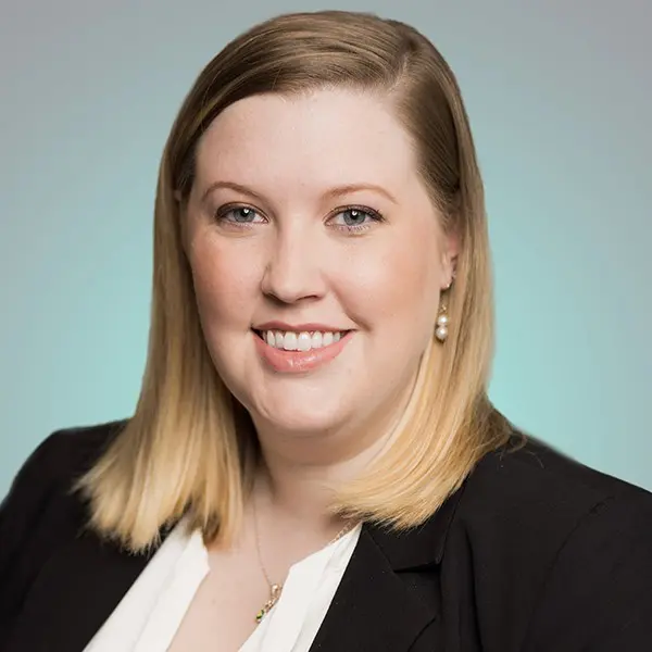Blonde woman smiling at camera, wearing a black blazer and white blouse; pearl earring and shoulder-length hair; soft teal gradient background.